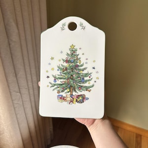 May include: White rectangular cutting board with a Christmas tree illustration. The tree is decorated with ornaments and topped with a star. Presents are at the base of the tree. The board has a hole for hanging.