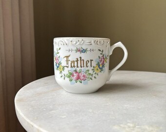 Peut inclure: Tasse en céramique blanche avec le mot "Father" en lettres dorées, entouré d'un motif floral de fleurs roses, bleues et jaunes. La tasse a une anse incurvée et un bord décoratif.