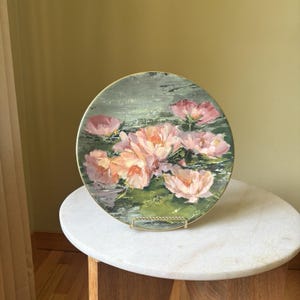 May include: A round decorative plate featuring an oil painting of soft pink flowers against a textured sage green and grey background. The plate has a thin gold rim and is displayed on a small gold wire stand, showcasing visible brushstrokes.