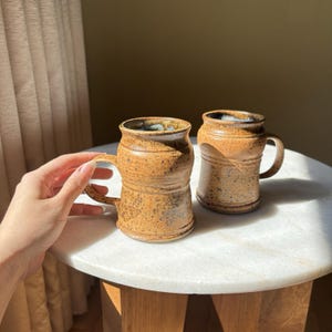May include: Two handmade, brown ceramic mugs with handles. The mugs have a textured surface and a slightly flared rim. The interior of the mugs has a blue-green glaze. The mugs are sitting on a white marble table.