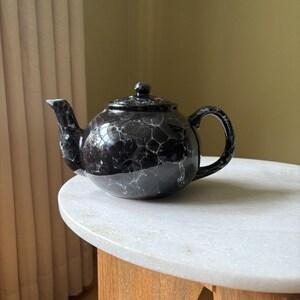 May include: A black ceramic teapot featuring a glossy finish with a white marbled or bubble-like pattern throughout the body, lid, and handle. The teapot has a rounded shape, a curved spout, and a small knob on the center of the lid.