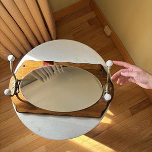 May include: A wall-mounted coat rack featuring a central oval mirror set into a rectangular wooden board with wavy edges. Two brass-toned metal hooks are attached to the sides, each topped with a small white spherical knob for hanging items.