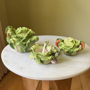 May include: A ceramic tea set featuring a teapot, a sugar bowl, and a creamer, all sculpted to resemble heads of green cabbage with textured leaves and small orange carrot accents on the handles and lids, arranged on a round white surface.