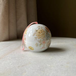 May include: A white porcelain ornament with a floral design in shades of yellow, pink, and blue. The ornament has a pink ribbon attached and is decorated with gold accents.