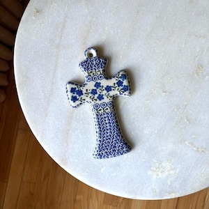 May include: A ceramic cross ornament featuring a white base with blue floral patterns, blue swirl accents, and a blue dotted texture. The cross has a small circular hole at the top for hanging and displays a detailed hand-painted aesthetic.
