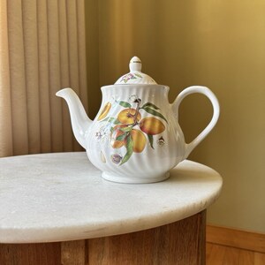 May include: A white porcelain teapot with a ribbed texture and a matching lid. The body features a colorful botanical illustration of orange and yellow apricots with green leaves and small white blossoms, accented by a small butterfly.
