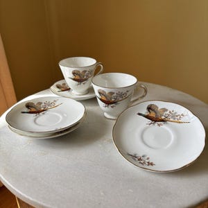 May include: White porcelain teacups and saucers with a pheasant design. The cups and saucers have a gold trim. The pheasant is depicted in shades of brown, green, and gold. The set includes two cups and three saucers.