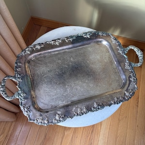 May include: A silver-toned rectangular serving tray with ornate floral detailing and handles. The tray has a decorative, textured border and a patterned interior. The handles are also detailed with floral designs. The tray is likely made of metal.