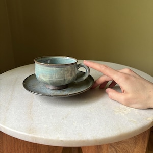 May include: A ceramic tea cup and matching saucer featuring a mottled glaze in shades of light blue, teal, and earthy brown. The cup has a rounded body with a small handle and a dark, unglazed base, while the saucer has a speckled finish.