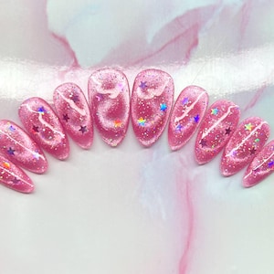 May include: Pink glitter nail set with star and swirl designs. The nails are oval-shaped and feature a shimmering, glittery finish. Each nail has a unique design with colorful stars and white swirls.
