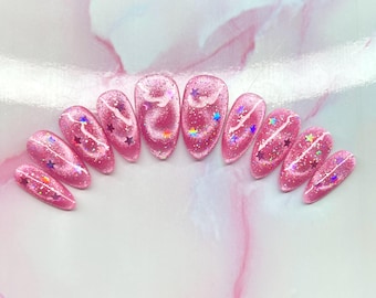May include: Pink glitter nail set with star and swirl designs. The nails are oval-shaped and feature a shimmering, glittery finish. Each nail has a unique design with colorful stars and white swirls.