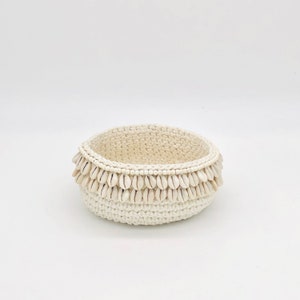 Puede incluir: Un cuenco de crochet blanco con un borde decorativo de conchas blancas. El cuenco es redondo y tiene una superficie lisa y texturizada.