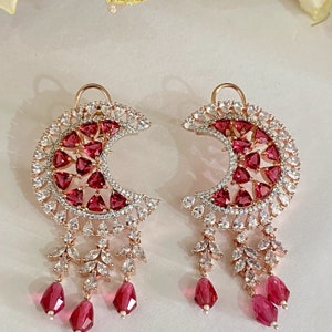May include: A pair of crescent moon shaped earrings with dangling charms. The earrings are made of rose gold and feature red and white cubic zirconia stones.