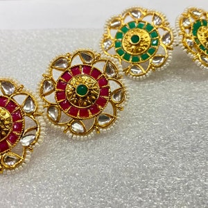 May include: Three pairs of gold-tone earrings with a circular design. Each earring features a central green stone surrounded by smaller pink or green stones. The earrings are accented with white pearls.