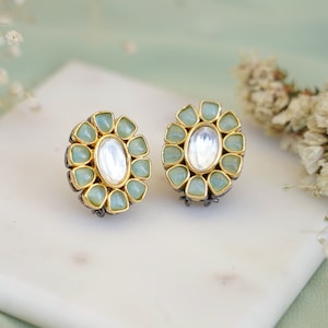 May include: A pair of gold and silver stud earrings with a floral design. The earrings have a central oval-shaped clear stone surrounded by smaller green stones.