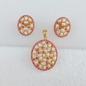 Könnte beinhalten: Ein goldfarbenes Schmuckset mit Anhänger und passenden Ohrringen. Der Anhänger und die Ohrringe sind rund, mit einem Rand aus kleinen roten Steinen. Die Mitte jedes Stücks ist mit weißen, unregelmäßig geformten Steinen gefüllt.