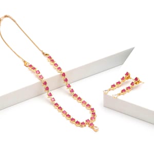 Puede incluir: Un conjunto de collar y pendientes colgantes en tono dorado, adornado con una serie de piedras redondas de color rosa vibrante. El collar incluye una cadena fina y una piedra transparente en forma de pera en el centro, a juego con los pendientes.