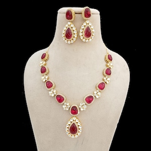 May include: A gold-coloured jewellery set comprising a necklace and matching earrings. The necklace and earrings feature teardrop-shaped red gemstones surrounded by small, clear stones. The necklace is displayed on a mannequin.