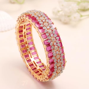 May include: A gold-toned bangle bracelet with a repeating pattern of rectangular pink stones. The bracelet is accented with clusters of clear and gold-toned stones, creating a floral design. The jewelry is set against a soft, neutral background.