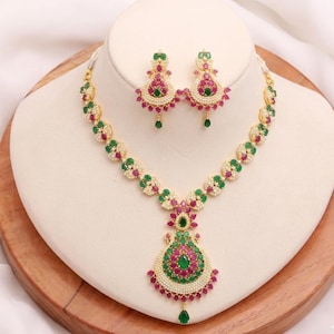May include: A gold-toned necklace and earring set featuring teardrop-shaped pendants and circular links. The jewelry is adorned with small white pearls, vibrant pink stones, and green stones arranged in a symmetrical, intricate floral-inspired pattern.