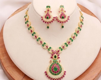 May include: A gold-toned necklace and earring set featuring teardrop-shaped pendants and circular links. The jewelry is adorned with small white pearls, vibrant pink stones, and green stones arranged in a symmetrical, intricate floral-inspired pattern.