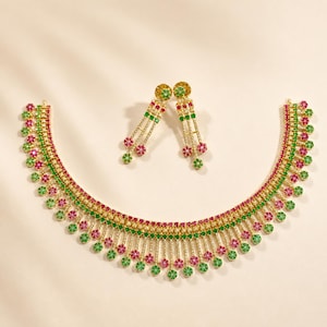 Op de afbeelding: Een goudkleurige ketting en bijpassende oorbellen set. De ketting en oorbellen hebben een ontwerp met rode, groene en goudkleurige stenen. De ketting heeft een gebogen vorm met bungelende accenten. De oorbellen hebben een bungelend ontwerp.