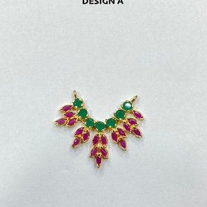 May include: A gold-toned jewelry connector labeled DESIGN A, featuring a curved arrangement of round green stones along the top and marquise-shaped magenta stones forming a leaf-like pattern extending downward from the center and sides.
