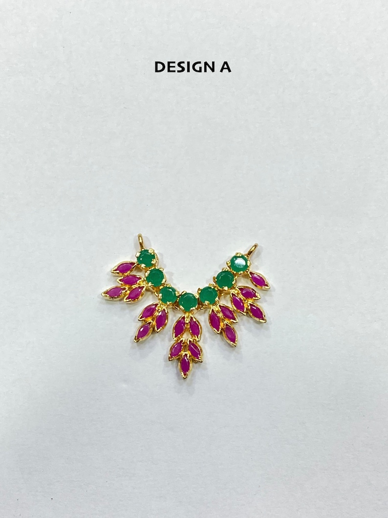 May include: A gold-toned jewelry connector labeled DESIGN A, featuring a curved arrangement of round green stones along the top and marquise-shaped magenta stones forming a leaf-like pattern extending downward from the center and sides.