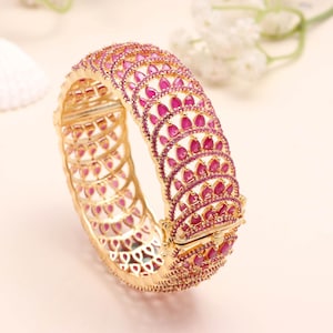 Op de afbeelding: Een goudkleurige armband met een reeks roze edelstenen. De edelstenen zijn in een herhalend patroon rond de armband gerangschikt. De armband heeft een sluiting en ingewikkelde details.