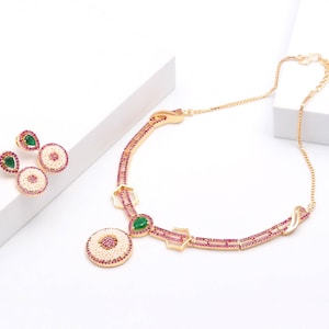 May include: A gold-toned jewelry set consisting of a necklace and matching earrings. The pieces feature small pink stones, white seed beads, and a central teardrop-shaped green stone. The necklace has a chain with a lobster claw clasp closure.