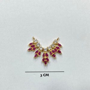 May include: A gold-toned jewelry connector labeled DESIGN D, featuring a fan-shaped arrangement of marquise-cut fuchsia stones and round clear stones. The piece measures 3 cm in width, as indicated by the text and line below the item.