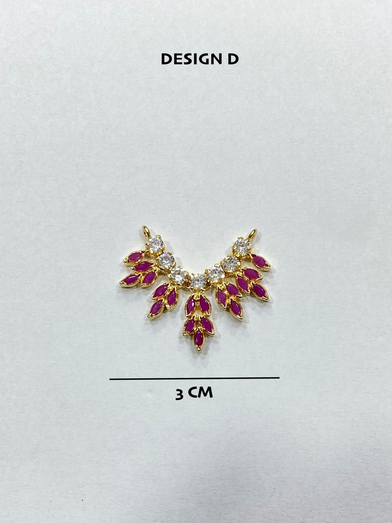 May include: A gold-toned jewelry connector labeled DESIGN D, featuring a fan-shaped arrangement of marquise-cut fuchsia stones and round clear stones. The piece measures 3 cm in width, as indicated by the text and line below the item.