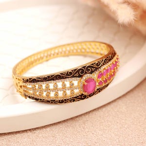 May include: A gold-colored bangle bracelet with intricate detailing. The bracelet features a central section with oval-shaped pink gemstones and smaller clear stones. The edges are adorned with black filigree patterns, creating a luxurious look.