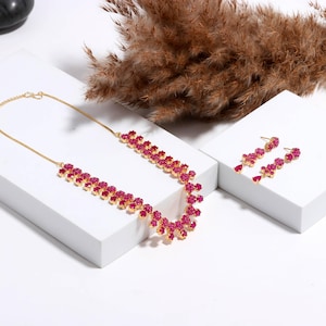 Op de afbeelding: Een goudkleurige ketting en bijpassende oorbellen set. De sieraden hebben een bloemmotief met levendige roze edelstenen. De ketting heeft een trapsgewijze opstelling en de oorbellen zijn van het druppeltype. De set wordt gepresenteerd op een wit oppervlak.