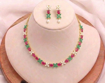 May include: A gold-toned necklace and matching earrings set. The necklace and earrings feature a floral design with red oval-shaped stones, green leaves, and white flower accents. The earrings have a single pearl drop.