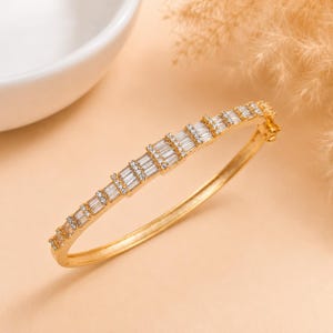 Può includere: Un bracciale rigido incernierato color oro con una fila di pietre trasparenti taglio baguette lungo la metà superiore. La fascia in metallo ha una finitura lucida e include un meccanismo di chiusura sicuro sul lato per l'apertura.
