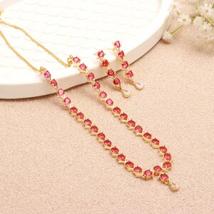 May include: A gold-toned jewelry set featuring a necklace and matching earrings. The necklace and earrings are adorned with vibrant pink gemstones. The earrings have a dangling teardrop-shaped accent. The set is displayed on a white tray and a light pink surface.