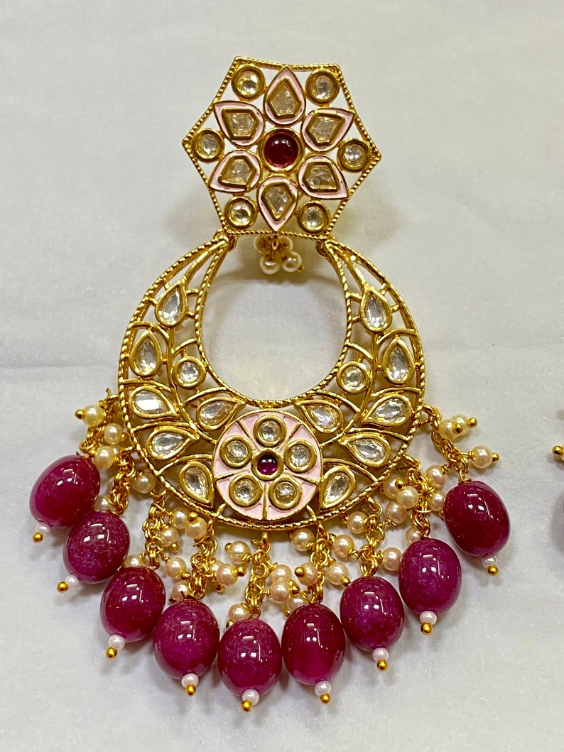 May include: Gold earrings with a pink and white enamel flower design, a crescent moon shape, and a dangling chain of burgundy beads.