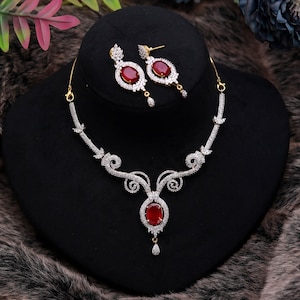 Puede incluir: Un deslumbrante conjunto de joyas con collar y pendientes a juego. El collar y los pendientes muestran una vibrante piedra preciosa ovalada roja rodeada de brillantes piedras transparentes. El conjunto se exhibe sobre un busto de joyería negro.