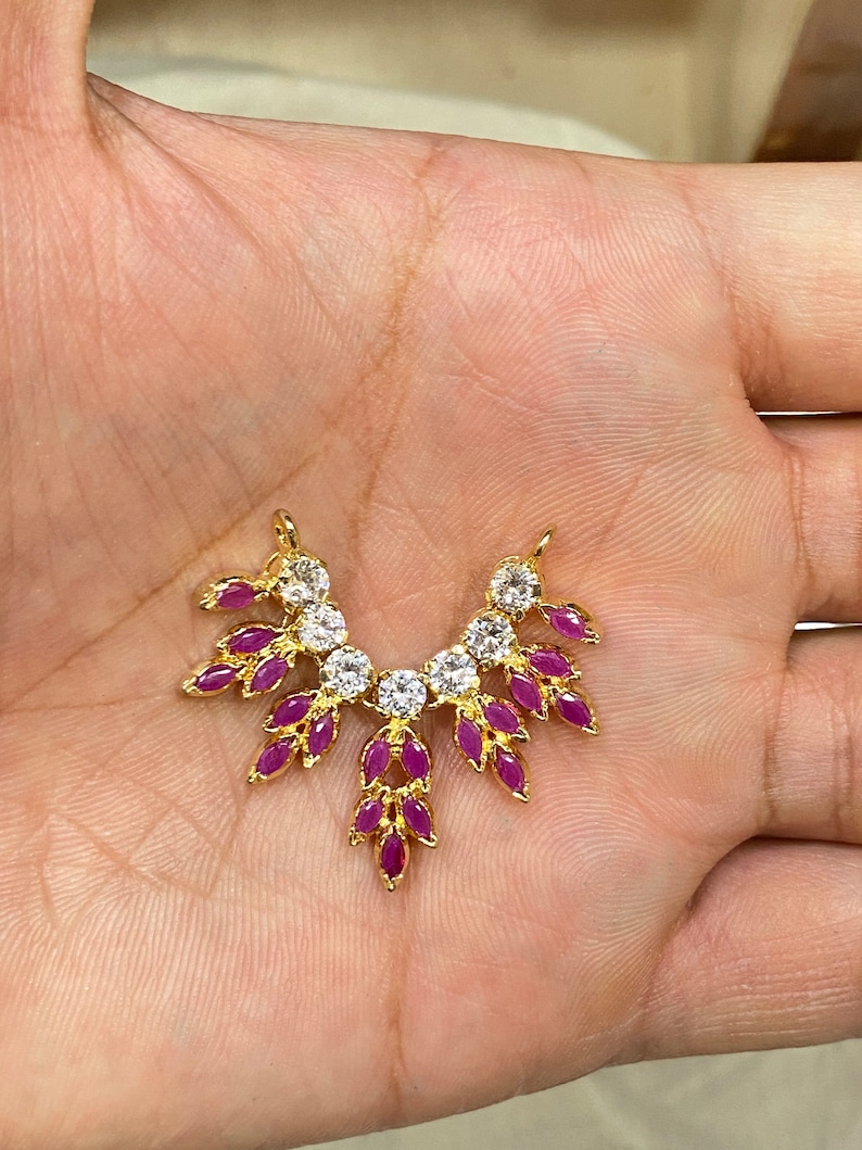 May include: A gold-toned jewelry connector featuring a fan-shaped arrangement of marquise-cut fuchsia stones and round clear stones. The piece has two small loops at the top for attachment, with a symmetrical design radiating from a central stone.