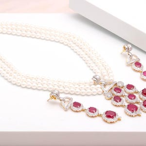 Puede incluir: Un collar de perlas de tres hilos con cierre dorado, combinado con pendientes a juego. Los pendientes y el colgante presentan piedras rojas de corte ovalado, rodeadas de pequeñas piedras transparentes, engastadas en oro. Las joyas se exhiben sobre una superficie blanca.