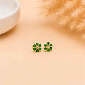 May include: A pair of gold stud earrings shaped like flowers, each featuring six teardrop-shaped green stones arranged around a central clear round stone. The earrings have a polished gold finish and a delicate, symmetrical floral design.