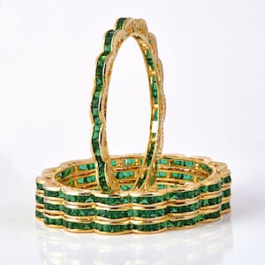 May include: A set of four gold bangles with green emerald-like stones. The bangles are stacked on top of each other.