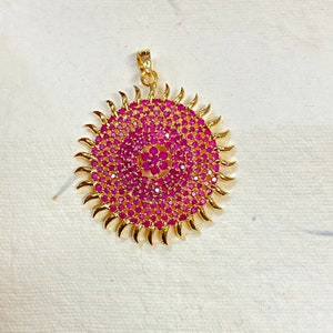Puede incluir: Un colgante de oro con un diseño circular que presenta un grupo central de piedras preciosas rosas rodeadas de piedras preciosas rosas más pequeñas. El colgante tiene un diseño de rayos de sol con rayos puntiagudos.