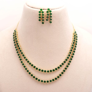 Puede incluir: Un conjunto de collar y pendientes dorados. El collar presenta dos hilos de pequeñas piedras redondas de color verde esmeralda. Los pendientes a juego tienen un diseño colgante con piedras verdes similares. Las joyas se exhiben en un maniquí blanco.