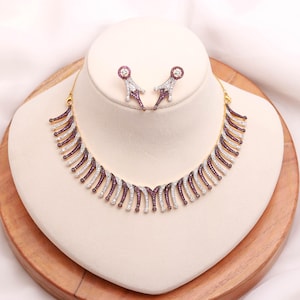 Puede incluir: Un collar dorado y un juego de pendientes a juego. El collar y los pendientes presentan un patrón repetido de piedras preciosas blancas y moradas alternas. Las joyas se exhiben sobre un busto blanco sobre un fondo de madera.