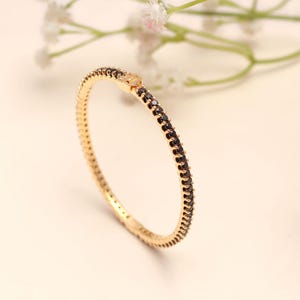 Könnte beinhalten: Ein goldfarbenes Armreif-Armband mit einer Reihe kleiner, schwarzer Edelsteine. Das Armband befindet sich vor einem weichen, neutralen Hintergrund mit zarten weißen Blüten.