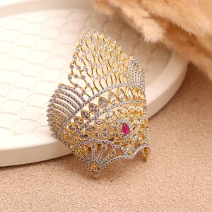 May include: A gold and silver-toned crown-shaped bracelet with clear and red gemstones. The bracelet features an intricate design with a central red gemstone and numerous clear stones, creating a sparkling effect. The jewelry piece is set against a neutral background.