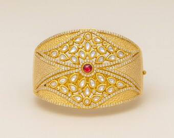 May include: A gold-toned cuff bracelet with a central design featuring a red gemstone surrounded by white teardrop-shaped stones. The bracelet has a textured band and is embellished with small, sparkling accents.