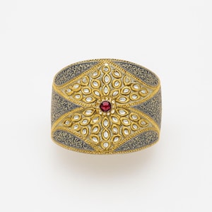 May include: A wide, ornate cuff bracelet with a central ruby-colored stone. The bracelet features gold-colored filigree and a dark gray, textured background. The design is symmetrical and detailed, showcasing a floral pattern.